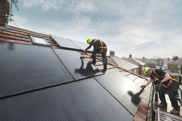 Two workers installing solar panels on a sloped roof, wearing safety harnesses and helmets