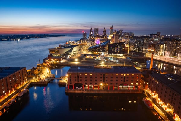 Aerial view of UK city waterfront at dusk, showing illuminated historic dock buildings, modern architecture, and the River