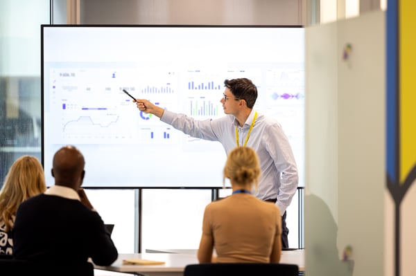 In a meeting room, a man gestures towards a graph displayed on a large TV screen.